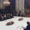George W. Bush delivering a speech behind the lectern at The Middle East Peace Conference at the Royal Palace in Madrid, Spain (30/10/1991). The numerous representatives surround a large rectangular white table, decorated with two flower arrangements and the signs of the participating countries: Israel, Lebanon, Syria, Egypt and Jordan-Palestine. In the background, journalists with cameras and several tapestries in an imposing white stone room. Two large lamps hang from the ceiling.