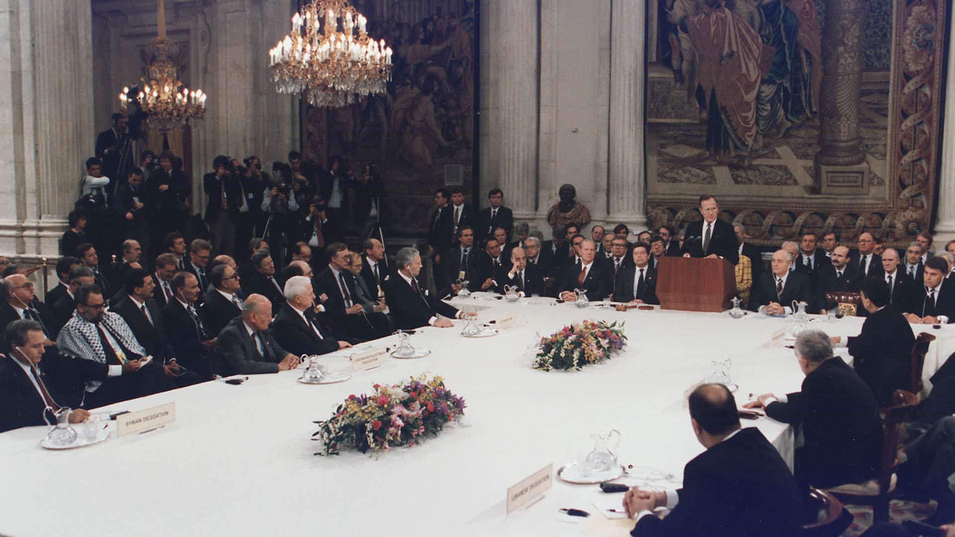 George W. Bush delivering a speech behind the lectern at The Middle East Peace Conference at the Royal Palace in Madrid, Spain (30/10/1991). The numerous representatives surround a large rectangular white table, decorated with two flower arrangements and the signs of the participating countries: Israel, Lebanon, Syria, Egypt and Jordan-Palestine. In the background, journalists with cameras and several tapestries in an imposing white stone room. Two large lamps hang from the ceiling.