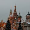 Vista de la fachada del Kremlin y algunos edificios circundantes cubiertos de nieve. Los edificios, predominantemente en tonos rojizos, destacan sobre el cielo grisáceo, casi de noche. Hay varios árboles y farolas que iluminan la escena.