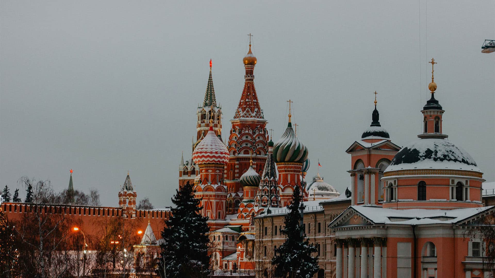 Vista de la fachada del Kremlin y algunos edificios circundantes cubiertos de nieve. Los edificios, predominantemente en tonos rojizos, destacan sobre el cielo grisáceo, casi de noche. Hay varios árboles y farolas que iluminan la escena.