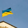 Una bandera de Ucrania corona el tejado de un edificio del gobierno. Sobre el cielo azul, destaca la parte superior del edificio, de color blanco y tejas marrones, y el mástil y la bandera ondeando.