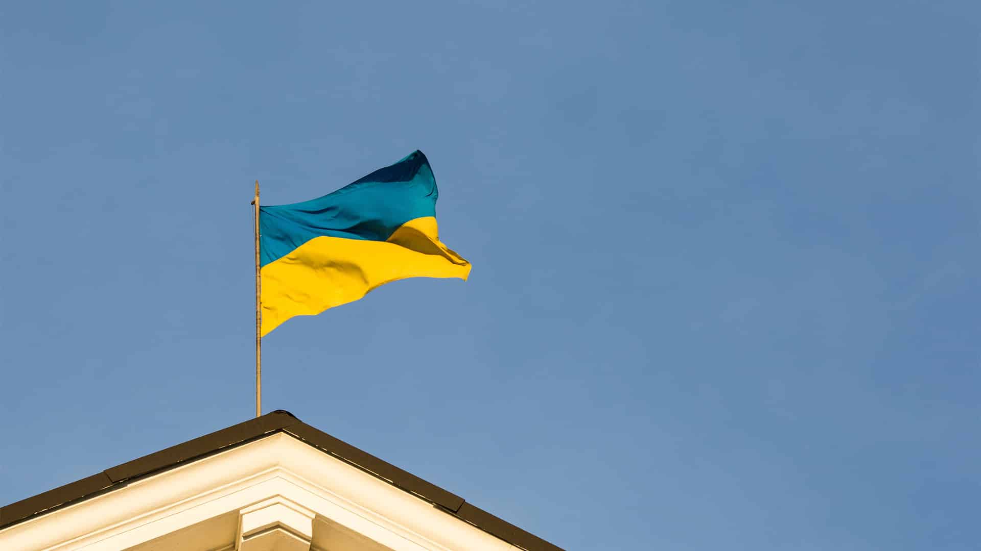 Una bandera de Ucrania corona el tejado de un edificio del gobierno. Sobre el cielo azul, destaca la parte superior del edificio, de color blanco y tejas marrones, y el mástil y la bandera ondeando.