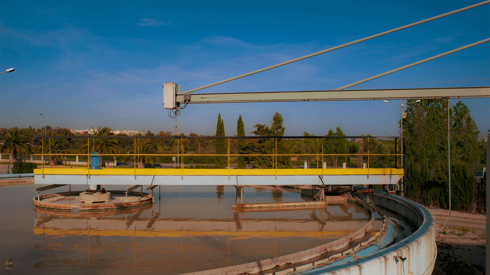 Estanque en una planta de tratamiento de agua en primer plano; al fondo, árboles desenfocados y una ciudad alejada, bajo un cielo azul despejado.