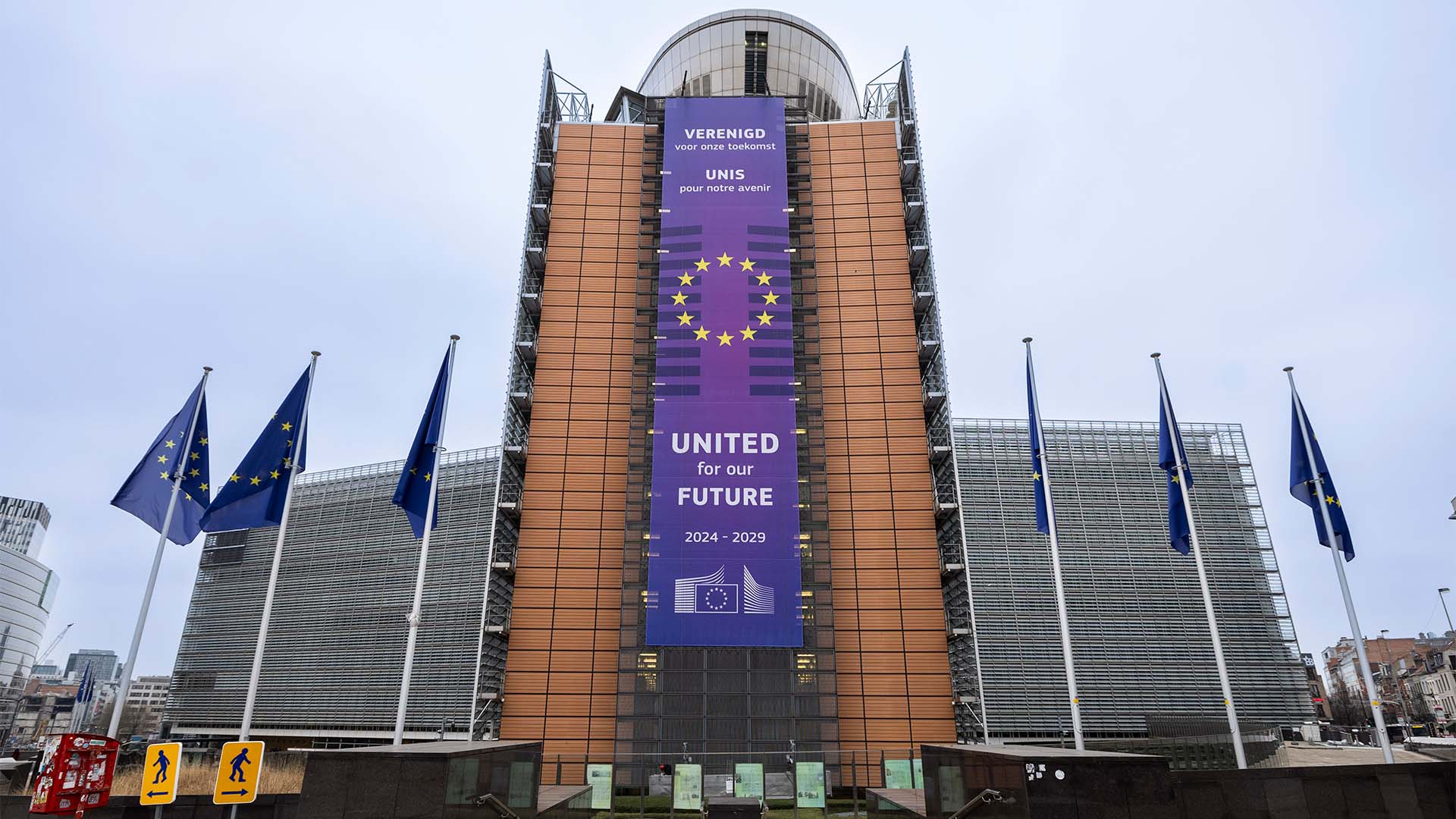 En el frente de la fachada color teja del Edificio Berlaymont, sede de la UE en Bruselas, un gran cartel con el texto “United for our future” y “2024-2029”, con el logo de la sede de la UE. A ambos lados, banderas de la UE y, más atrás, la forma asimétrica de dos de las alas del edificio, de aspecto moderno, con cristal y metal. La imagen fue tomada en la durante el despliegue de la pancarta, en diciembre de 2024.