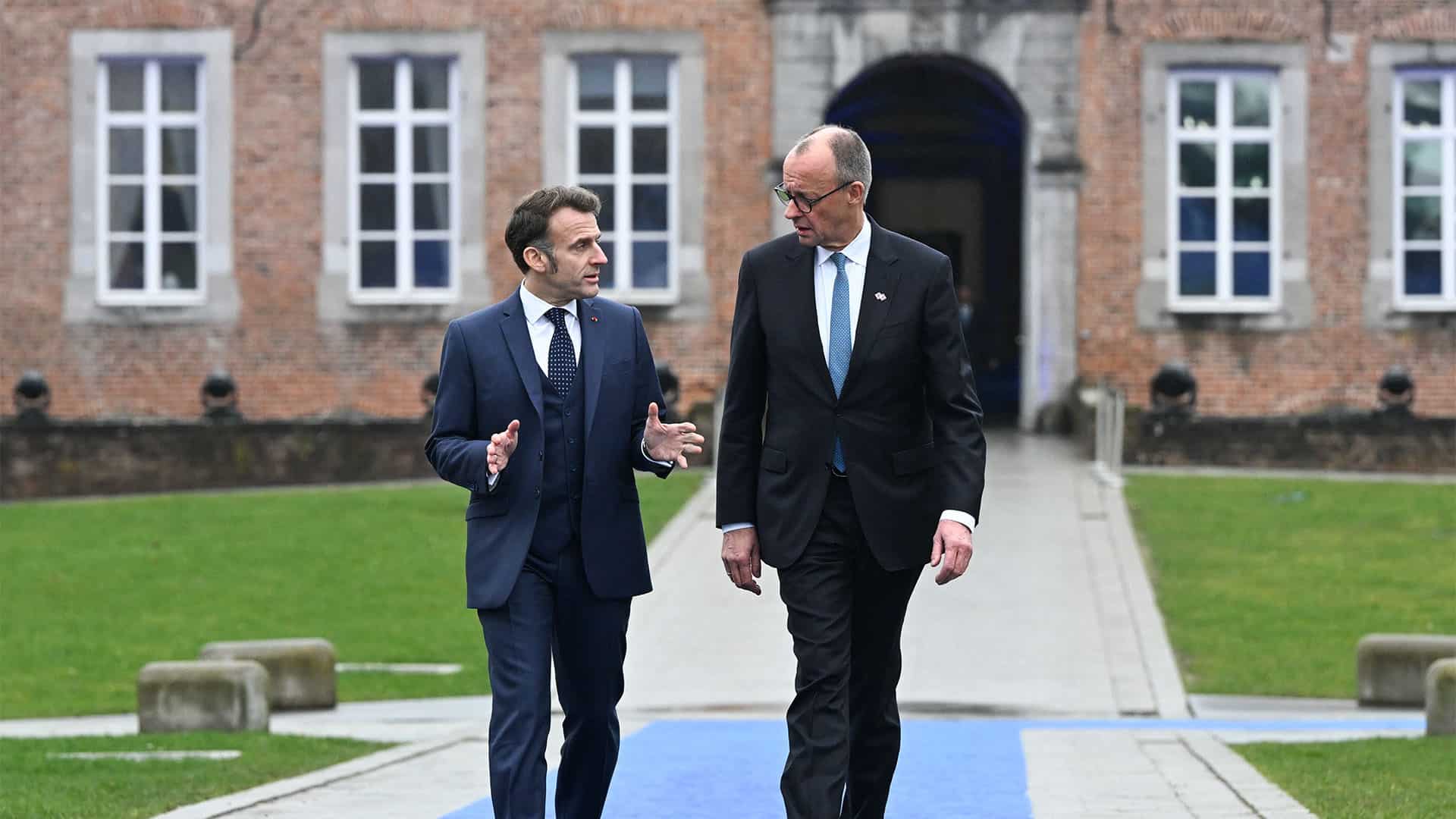 Emmanuel Macron, presidente de Francia, y Friedrich Merz, canciller de Alemania, hablan en la llegada a la reunión informal de los miembros del Consejo Europeo en el castillo de Alden Biesen, en Bélgica. En primer plano, ambos representantes, que caminan por el paseo de acceso, rodeado del verde del césped, y, al fondo, una de las fachadas del castillo, que mezcla el color teja con el gris de las piedras, con una entrada en arco, y varias ventanas.