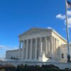 Vista lateral de la sede del Tribunal Supremo de EEUU en Washington D.C. El edificio, predominantemente blanco y con un marcado estilo neoclásico, destaca en lo alto. A la derecha de la escalinata de entrada, un gran mástil con la bandera de EEUU ondeando. El cielo está muy despejado y es de un color azul intenso.