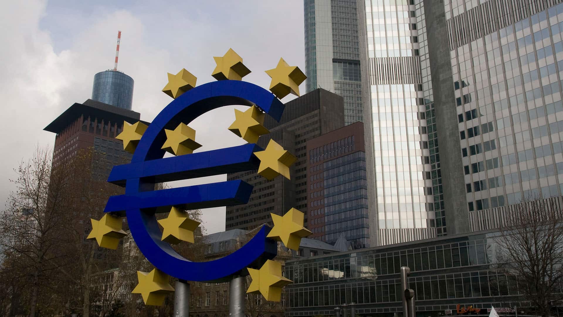 Icono del euro delante de la sede del Banco Central Europeo, en Frankfurt. El icono, de grandes dimensiones, en primer plano, deja detrás el edificio acristalado y metálico del Banco Central Europeo. Al lado del edificio, otros de color teja.