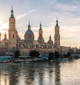 View of the Basilica del Pilar in Zaragoza from the River Ebro, which runs through the city. The cathedral stands out against the blue and yellow sky at sunset. In the background, part of the city and one of the bridges that cross it.