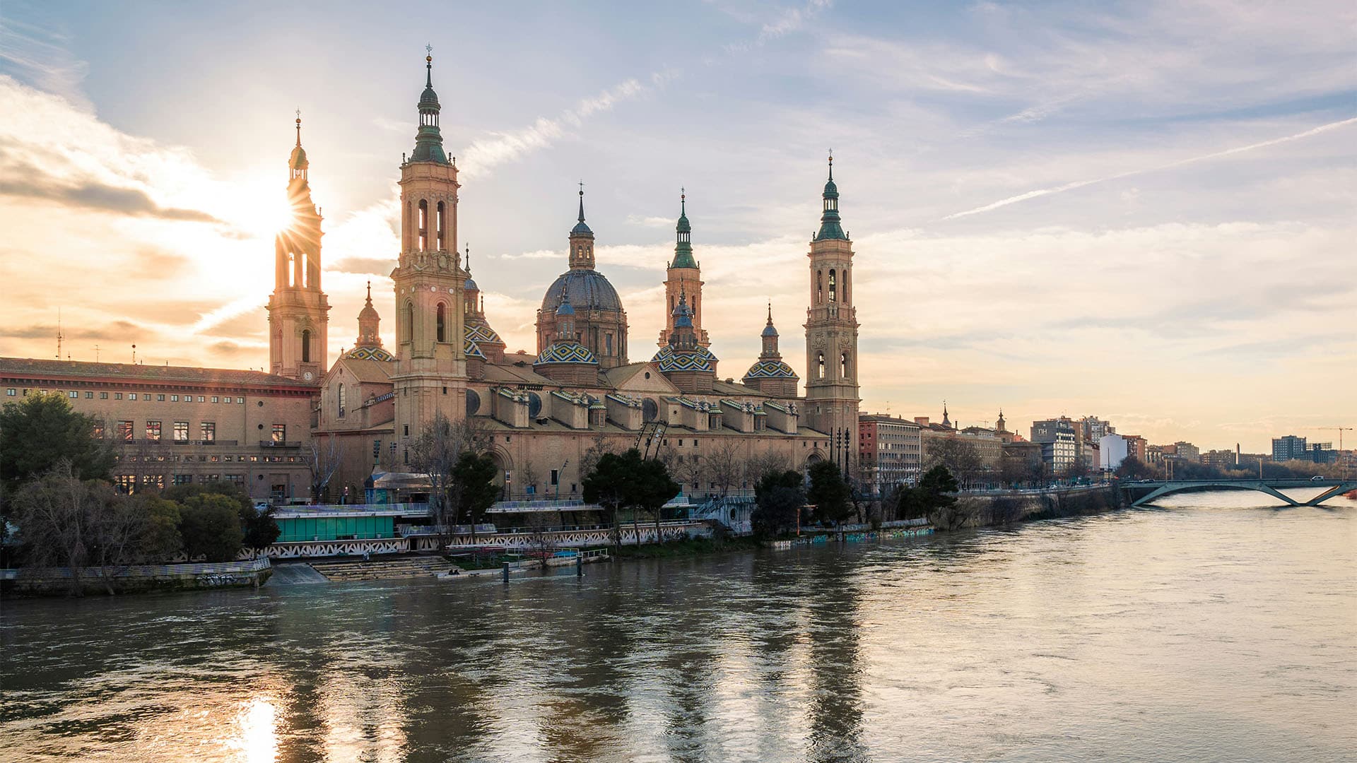 View of the Basilica del Pilar in Zaragoza from the River Ebro, which runs through the city. The cathedral stands out against the blue and yellow sky at sunset. In the background, part of the city and one of the bridges that cross it.