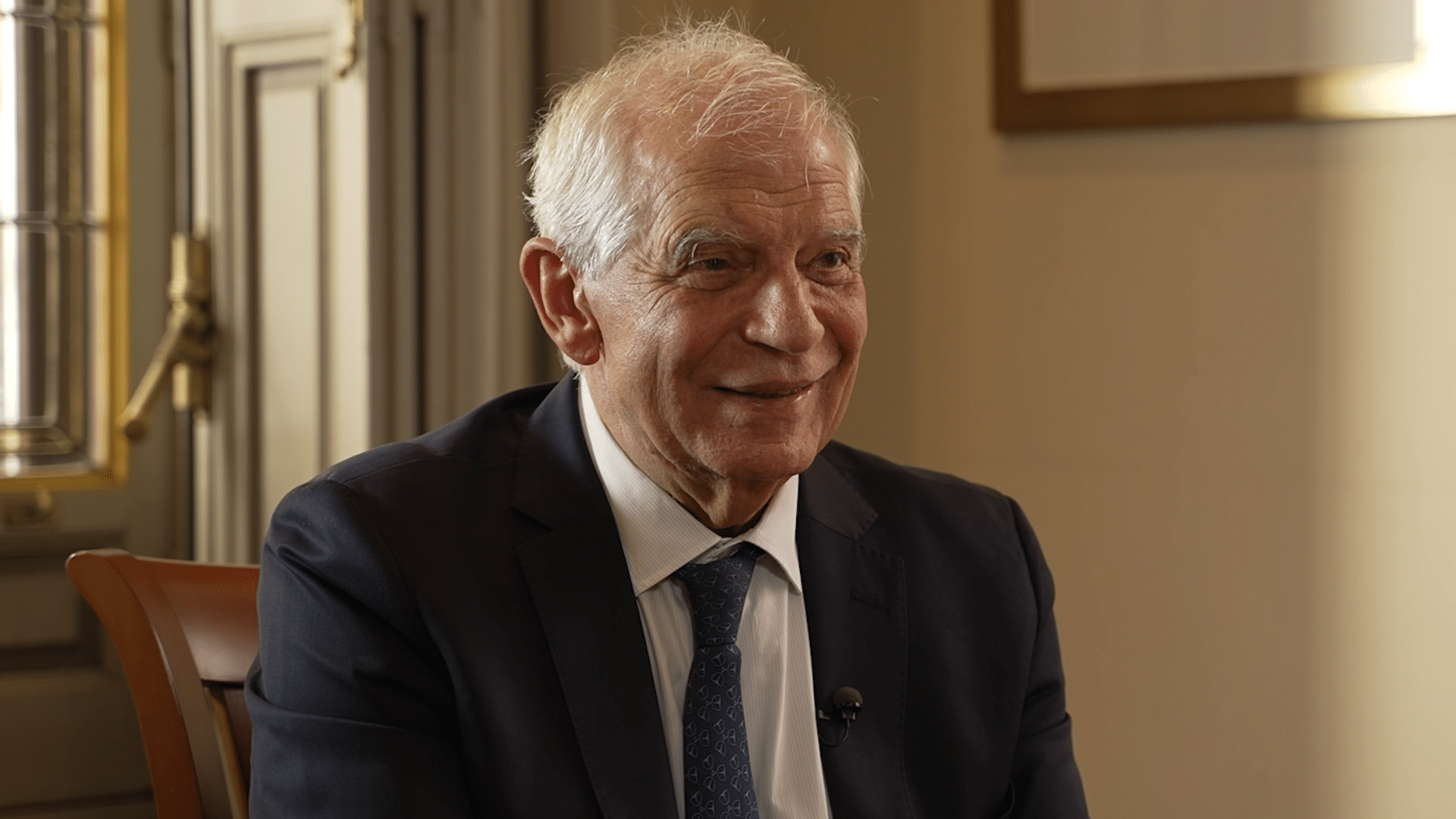 Josep Borrell sonríe en la grabación del vídeo por los 40 años de España en la Unión Europea en la sede del Real Instituto Elcano. Borrell está sentado delante de una ventana y al lado de un marco de color dorado, que ocupan parcialmente la imagen.