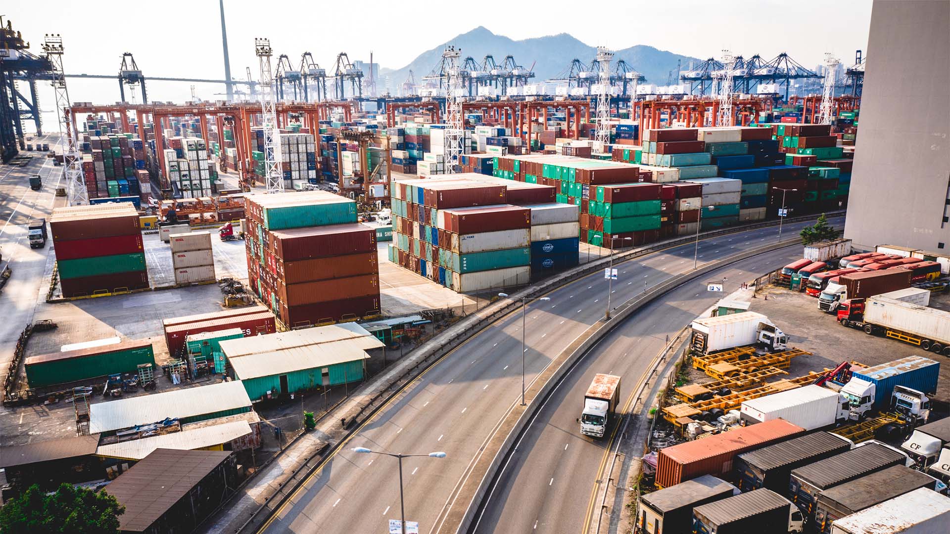 Zona portuaria en Kowloon, Hong Kong, con numerosos contenedores apilados en distintos colores y grúas industriales al fondo. En primer plano, una autopista curva casi vacía con algunos camiones circulando, junto a áreas logísticas y vehículos estacionados. En la distancia se distinguen montañas y estructuras portuarias.
