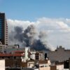 Una densa columna de humo gris oscuro y negro asciende verticalmente desde una zona situada detrás de varios edificios residenciales. En primer plano se observan azoteas y cubiertas de diferentes alturas, entre las que destaca un edificio más alto de color marrón con múltiples ventanas. El humo contrasta con un cielo nublado pero luminoso.