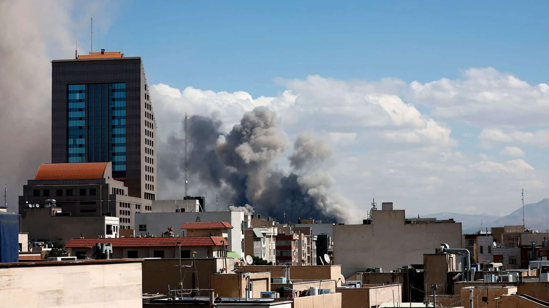 Una densa columna de humo gris oscuro y negro asciende verticalmente desde una zona situada detrás de varios edificios residenciales. En primer plano se observan azoteas y cubiertas de diferentes alturas, entre las que destaca un edificio más alto de color marrón con múltiples ventanas. El humo contrasta con un cielo nublado pero luminoso.