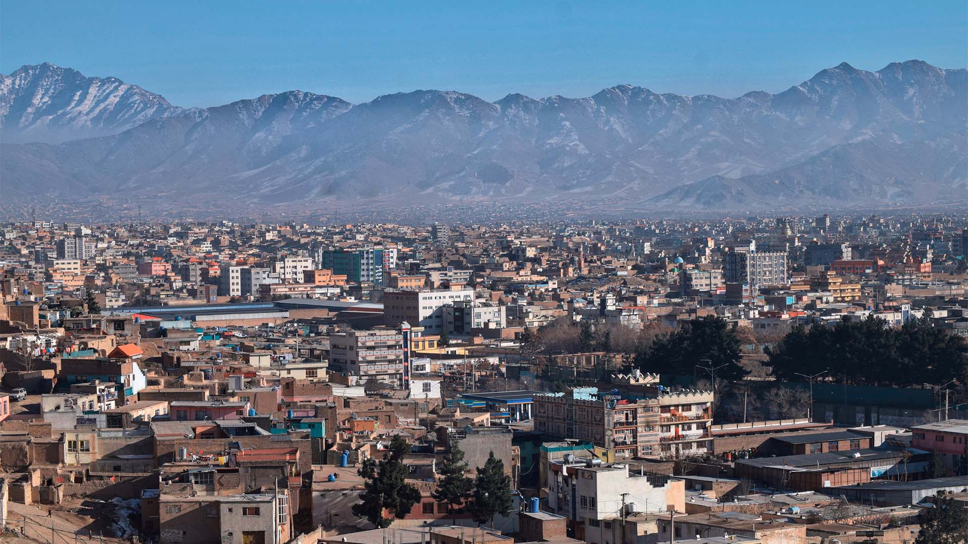 Vista general de la ciudad de Kabul, Afganistán. En primer plano, multitud de edificios residenciales y casas de baja altura, y, al fondo, difuminadas, las montañas de la ciudad delante de un cielo azul muy claro y despejado.