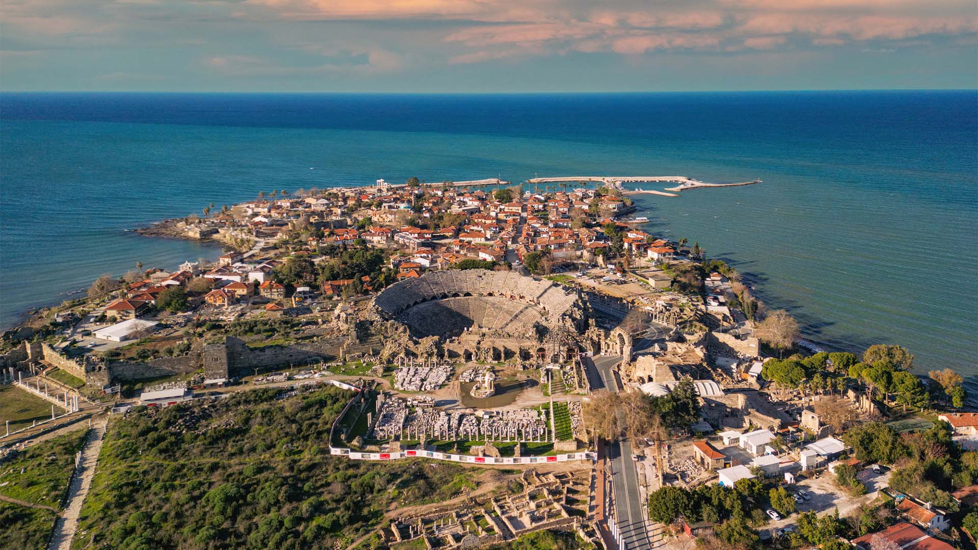 Vista aérea del casco antiguo de Antalya, Turquía, próxima sede de la COP 31. Destaca, en el centro de la imagen, el teatro romano de Aspendos, rodeado de algunas de las ruinas romanas aún conservadas en la ciudad. Detrás, edificios residenciales y, al fondo, parte del puerto y el mar abierto de un color azul intenso. El cielo es claro, aunque algo nublado.