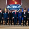 Doce representantes de los países latinoamericanos que integran Shield of the Americas posan junto al presidente de Estados Unidos en un podio de dos alturas, durante un acto oficial en el Hotel Trump National Doral, en Miami. Todos están de pie, formando dos filas ordenadas. Detrás de ellos se alinean las trece banderas de los países firmantes. En el centro del fondo destaca el logotipo de ‘Shield of the Americas’, un gran escudo de color rojo predominante que incluye el mapa del continente americano y las palabras ‘Shield of the Americas’ y ‘Doral 2026’ en letras grandes. A ambos lados del escenario cuelgan cortinas rojas.