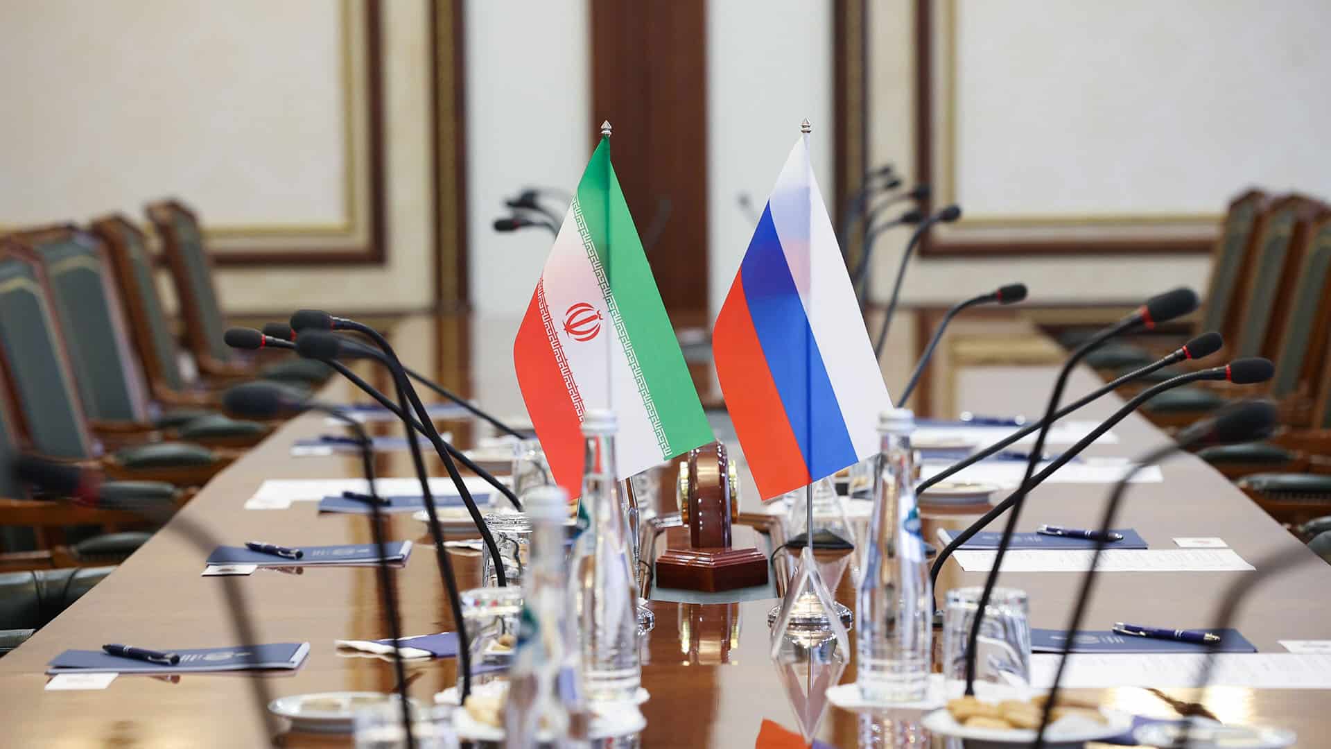 Detalle de una mesa de reunión formal con dos pequeñas banderas de Irán y Rusia colocadas en el centro rodeada de micrófonos, documentos y botellas de agua.