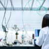 Female scientist in a white lab coat conducting an experiment at a laboratory bench, surrounded by test tubes, measurement equipment, and cables, while observing a results screen beneath a metal structure.