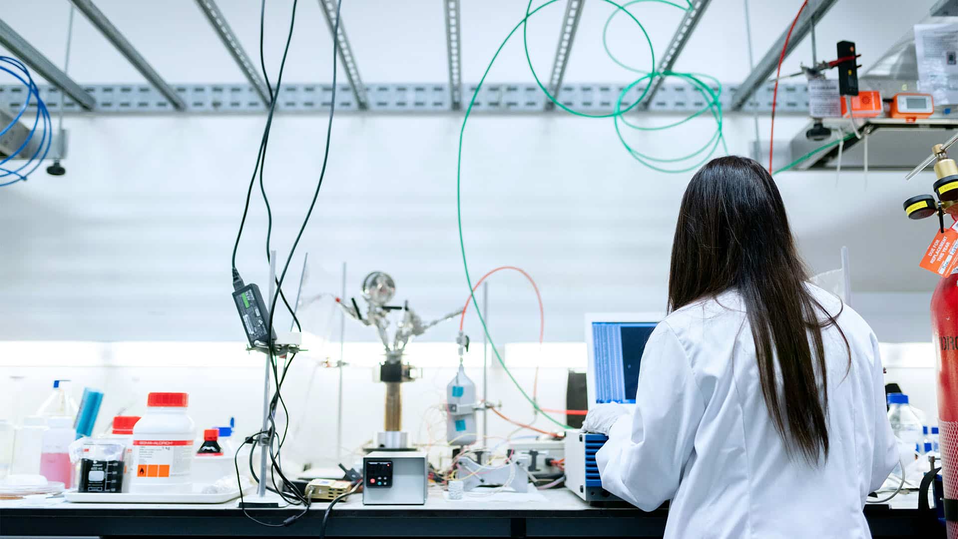 Female scientist in a white lab coat conducting an experiment at a laboratory bench, surrounded by test tubes, measurement equipment, and cables, while observing a results screen beneath a metal structure.