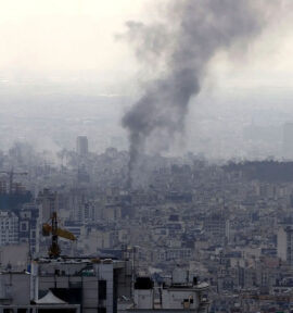 Vista panorámica de Teherán mientras una espesa columna de humo negro se eleva desde la ciudad tras una explosión, entre densos edificios y grúas de construcción bajo un cielo grisáceo.