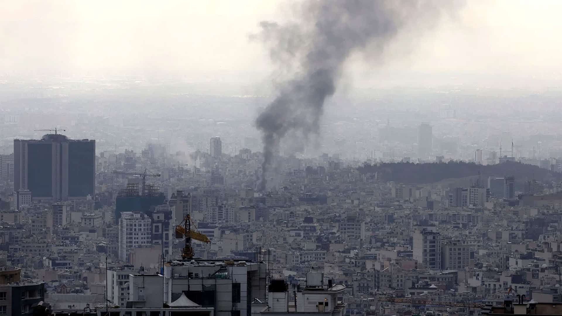 Panoramic view of Tehran as a thick column of black smoke rises from the city after an explosion, among dense buildings and construction cranes under a greyish sky.
