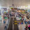 The interior of Malaga Airport, bustling with travellers. People walk in both directions and others wait in line to board. In the centre there are moving walkways and, on both sides, boarding gates and some shops. The space is large, white and with large windows that let in natural light.
