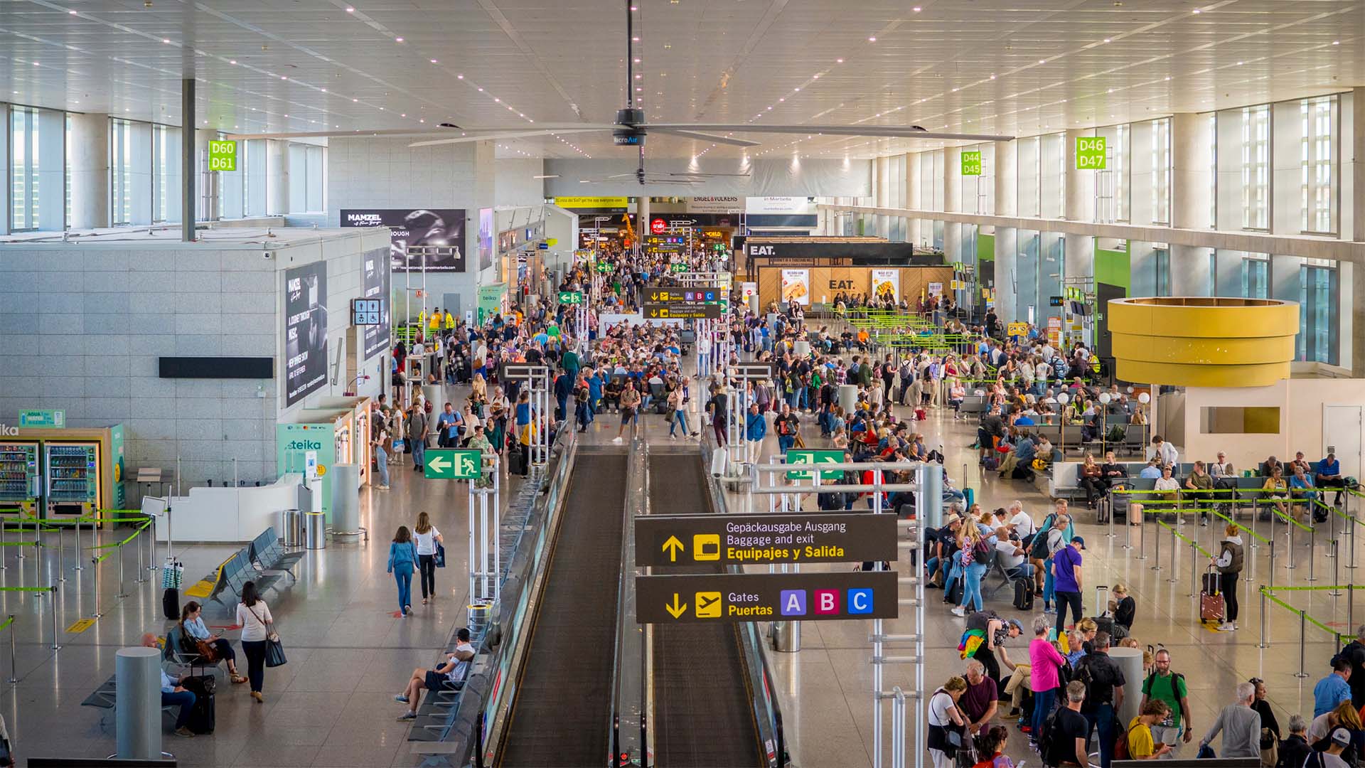 The interior of Malaga Airport, bustling with travellers. People walk in both directions and others wait in line to board. In the centre there are moving walkways and, on both sides, boarding gates and some shops. The space is large, white and with large windows that let in natural light.