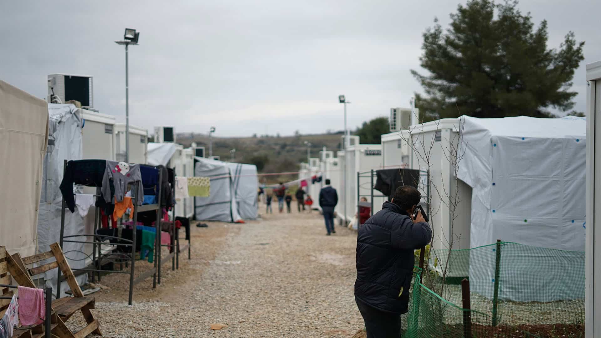 Hombre en primer plano hablando por teléfono en un albergue, con casetas blancas alineadas a ambos lados de un camino de grava y estructuras metálicas con ropa tendida a la izquierda; al fondo, varias personas caminan bajo un cielo muy nublado.