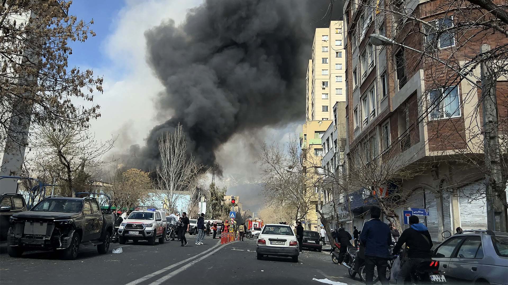 Una densa columna de humo negro se eleva desde el fondo de una calle en una ciudad, proveniente de una explosión reciente. En primer plano, vehículos detenidos y personas observan la escena. Los edificios residenciales flanquean la vía y el ambiente refleja tensión y emergencia tras el suceso.