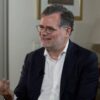 Wolfgang Schmidth conversa sonriendo y enfatizando con sus manos en la grabación de su entrevista en la sede del Real Instituto Elcano. Schmidt está sentado cerca de una ventana y al lado de un marco de color dorado y una lámpara blanca y transparente, que ocupan parcialmente el fondo de la imagen.