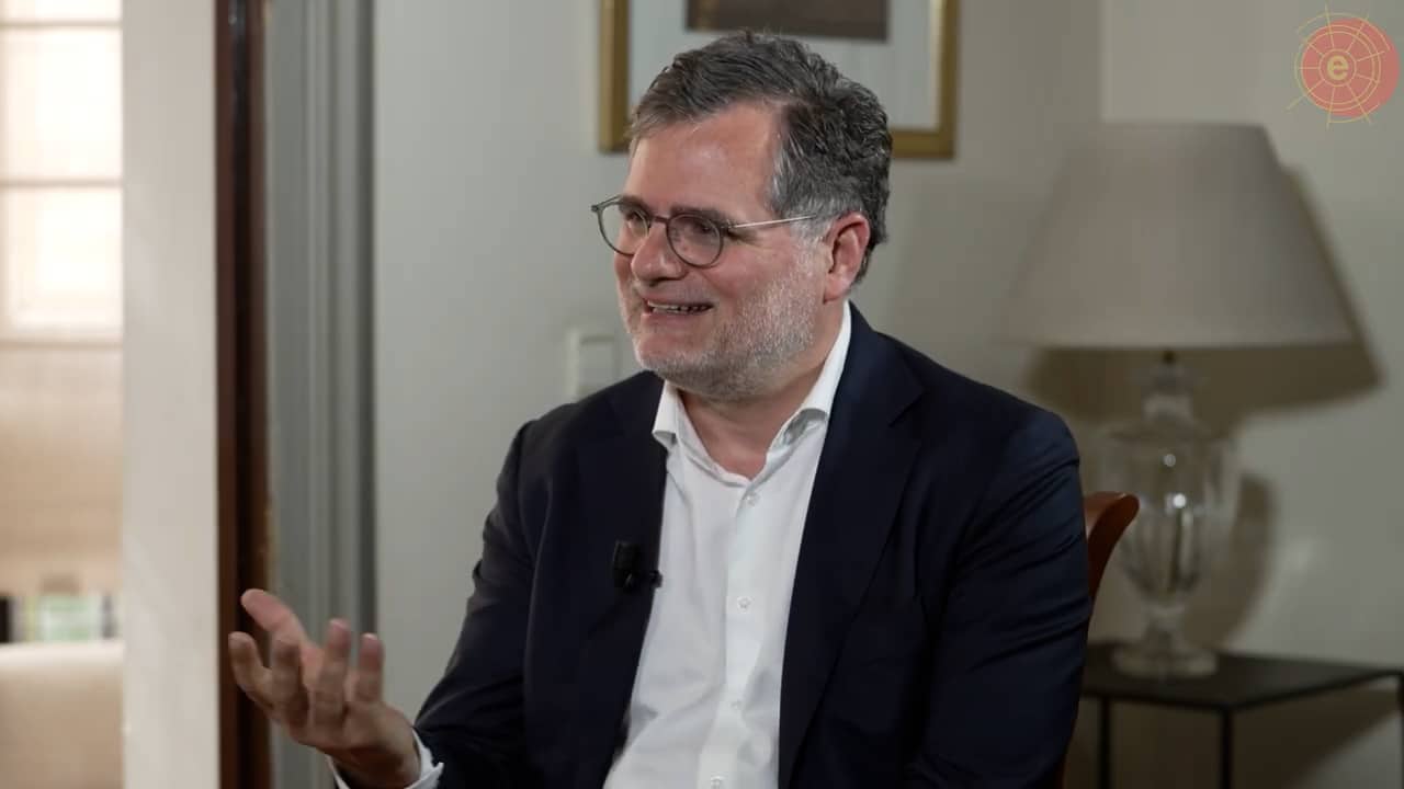 Wolfgang Schmidth conversa sonriendo y enfatizando con sus manos en la grabación de su entrevista en la sede del Real Instituto Elcano. Schmidt está sentado cerca de una ventana y al lado de un marco de color dorado y una lámpara blanca y transparente, que ocupan parcialmente el fondo de la imagen.