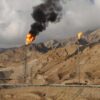 Paisaje de las instalaciones del yacimiento de gas de South Pars en Irán. En primer plano, una tubería de gas natural sobre pilares, un montículos de tubos, y varios containers de colores apilados. Detrás, dos torres de gas encendidas, la primera con una llamarada grande desprendiendo una columna de humo oscura y la segunda con una llamarada más pequeña. Al fondo, el paisaje de las montañas desérticas con el cielo nublado.