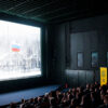 Sala de cine durante la proyección del documental Mr. Nobody Against Putin en el festival Artdocfest de Riga, Letonia. A la izquierda, una pantalla donde se proyecta un fragmento del documental, una escena invernal con una bandera rusa ondeando. A la derecha, el público observa desde sus asientos en la oscuridad; Entre los asientos, se distingue un cartel amarillo con el nombre del festival.