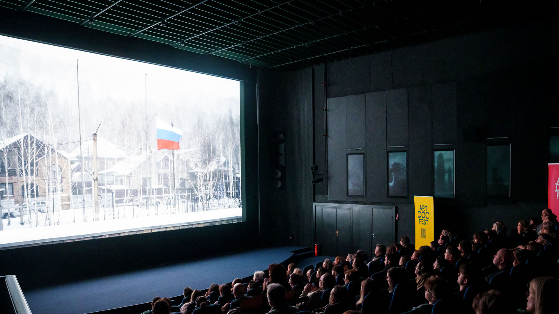 Sala de cine durante la proyección del documental Mr. Nobody Against Putin en el festival Artdocfest de Riga, Letonia. A la izquierda, una pantalla donde se proyecta un fragmento del documental, una escena invernal con una bandera rusa ondeando. A la derecha, el público observa desde sus asientos en la oscuridad; Entre los asientos, se distingue un cartel amarillo con el nombre del festival.