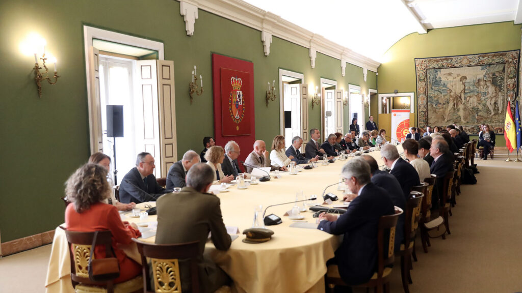 Vista en diagonal de la sala de la reunión de los miembros Consejo Científico del Real Instituto Elcano presidida por S.M. el Rey. Los miembros están sentados en una mesa alargada. Al final a la derecha, hay otro grupo de personas sentadas frente a un tapiz que cuelga en la pared del fondo, y a un lado las banderas de España y la UE. 