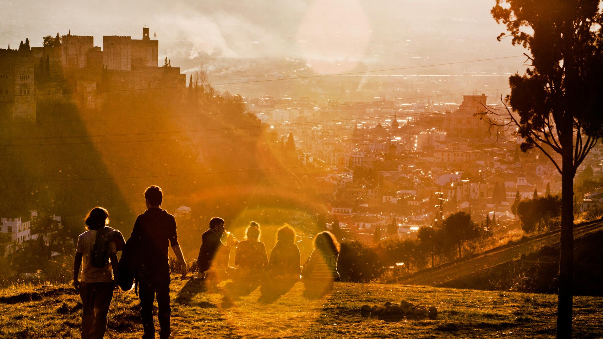 Parejas y personas en una colina observado la puesta de sol en Granada (España). A la derecha un árbol, y al fondo se divisa el paisaje de la ciudad.