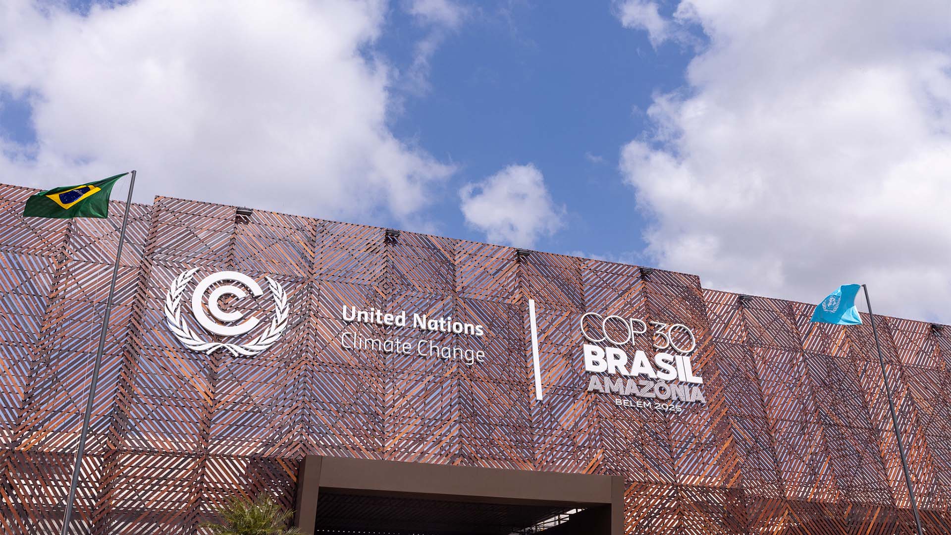 The modern COP30 building entrance in Belem, Brazil, featuring a bronze-colored metal latticework. On the facade, the flags of Brazil and the United Nations stand out against the brown-toned design. The United Nations Climate Change logo is displayed, along with the words “United Nations Climate Change” and the phrase "COP30 Brazil Amazonia Belem 2025".
