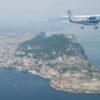 Aerial view of the Rock of Gibraltar surrounded by the sea, with the town, the port and the airport runway visible, whilst a light aircraft flies over the territory in a clear sky.