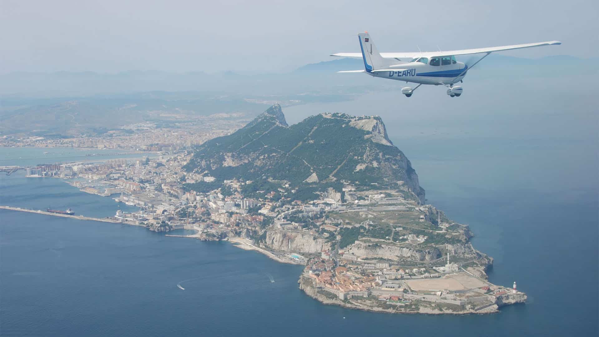 Aerial view of the Rock of Gibraltar surrounded by the sea, with the town, the port and the airport runway visible, whilst a light aircraft flies over the territory in a clear sky.