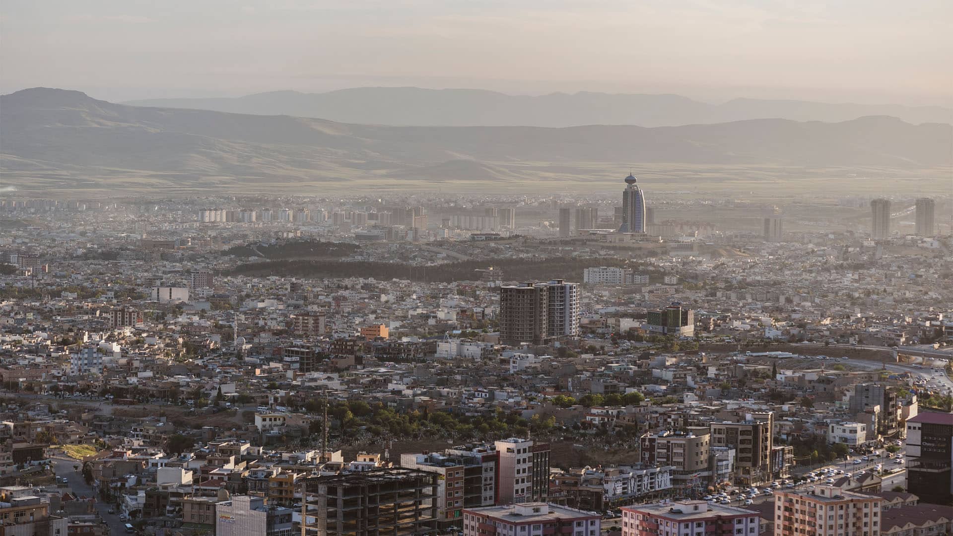 Panoramic view of the city of Sulaymaniyah, in Iraqi Kurdistan, with dense buildings in the foreground and mountains in the background under soft light.