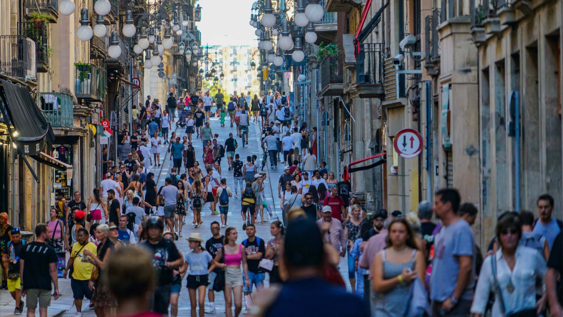 Calle estrecha del casco antiguo de Barcelona con arquitectura tradicional, farolas, balcones y tiendas, llena de personas en un día de verano.