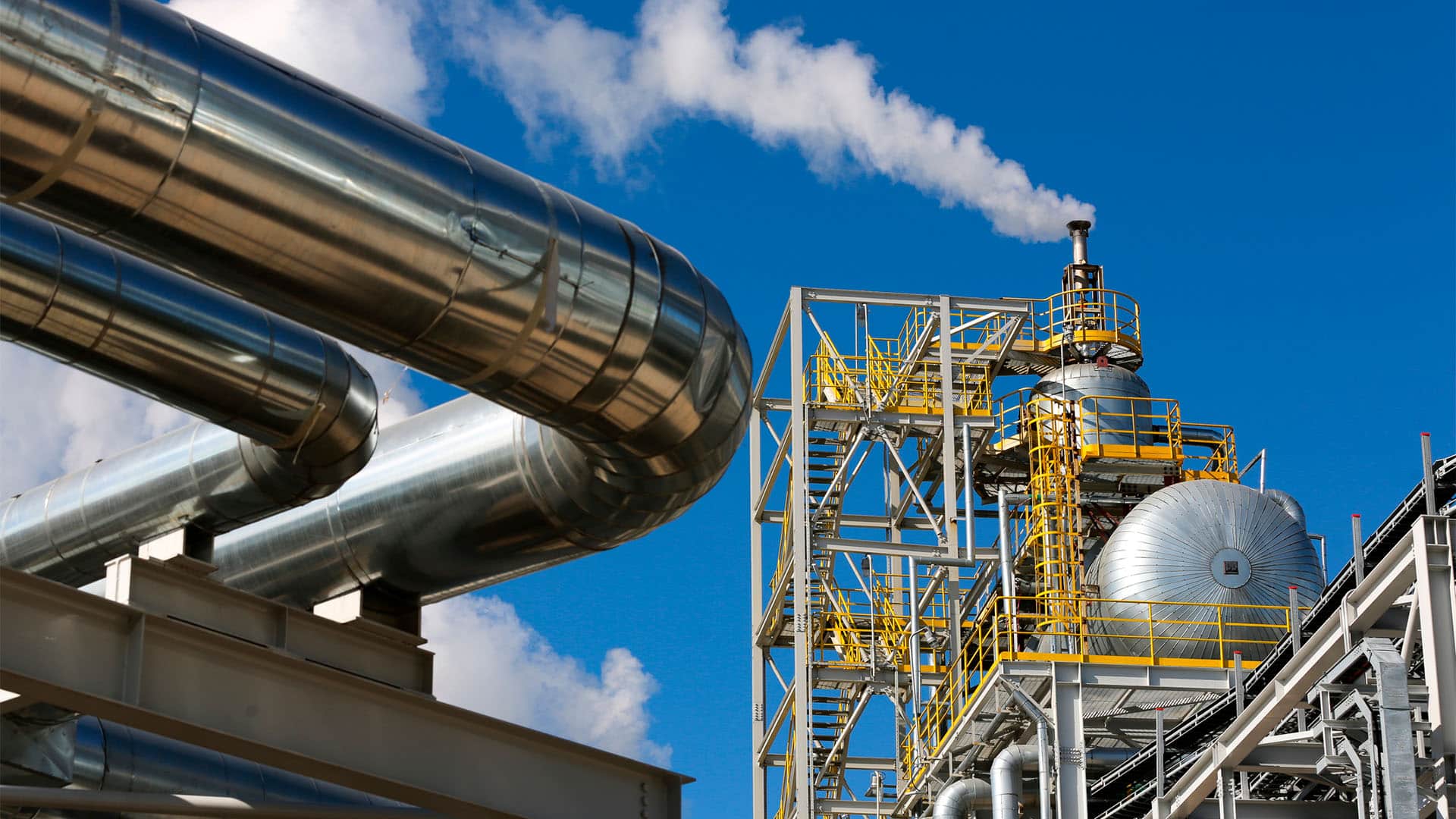 Instalación industrial con grandes tuberías metálicas y una torre de producción de amoníaco; desde la parte superior se eleva vapor blanco hacia el cielo azul.