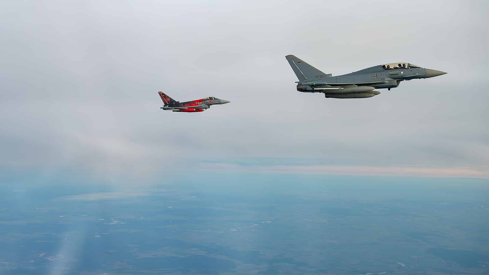 Dos aviones de combate Eurofighter Typhoon de la Fuerza Aérea alemana vuelan en formación junto a un bombardero B-52H Stratofortress de la Fuerza Aérea de Estados Unidos durante una misión en Europa. Los aviones sobrevuelan un cielo nublado y, debajo, se distinguen extensas áreas de terreno.