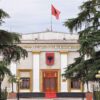 Fachada del Parlamento de Albania, con columnas blancas, escudo nacional y bandera albanesa en lo alto, rodeado de árboles y una entrada con escaleras y alfombra roja. El cielo es claro, aunque nublado.