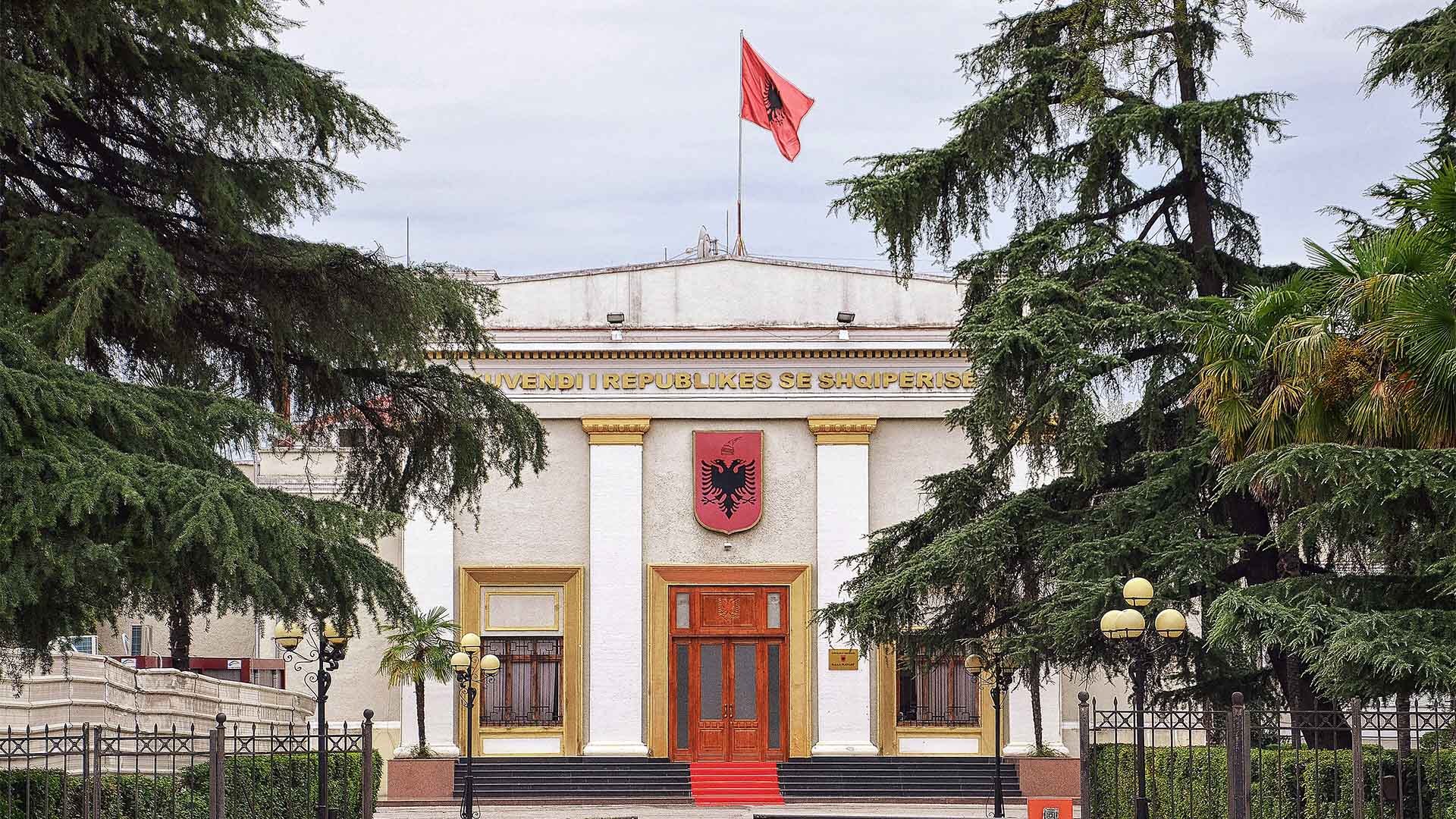 Fachada del Parlamento de Albania, con columnas blancas, escudo nacional y bandera albanesa en lo alto, rodeado de árboles y una entrada con escaleras y alfombra roja. El cielo es claro, aunque nublado.