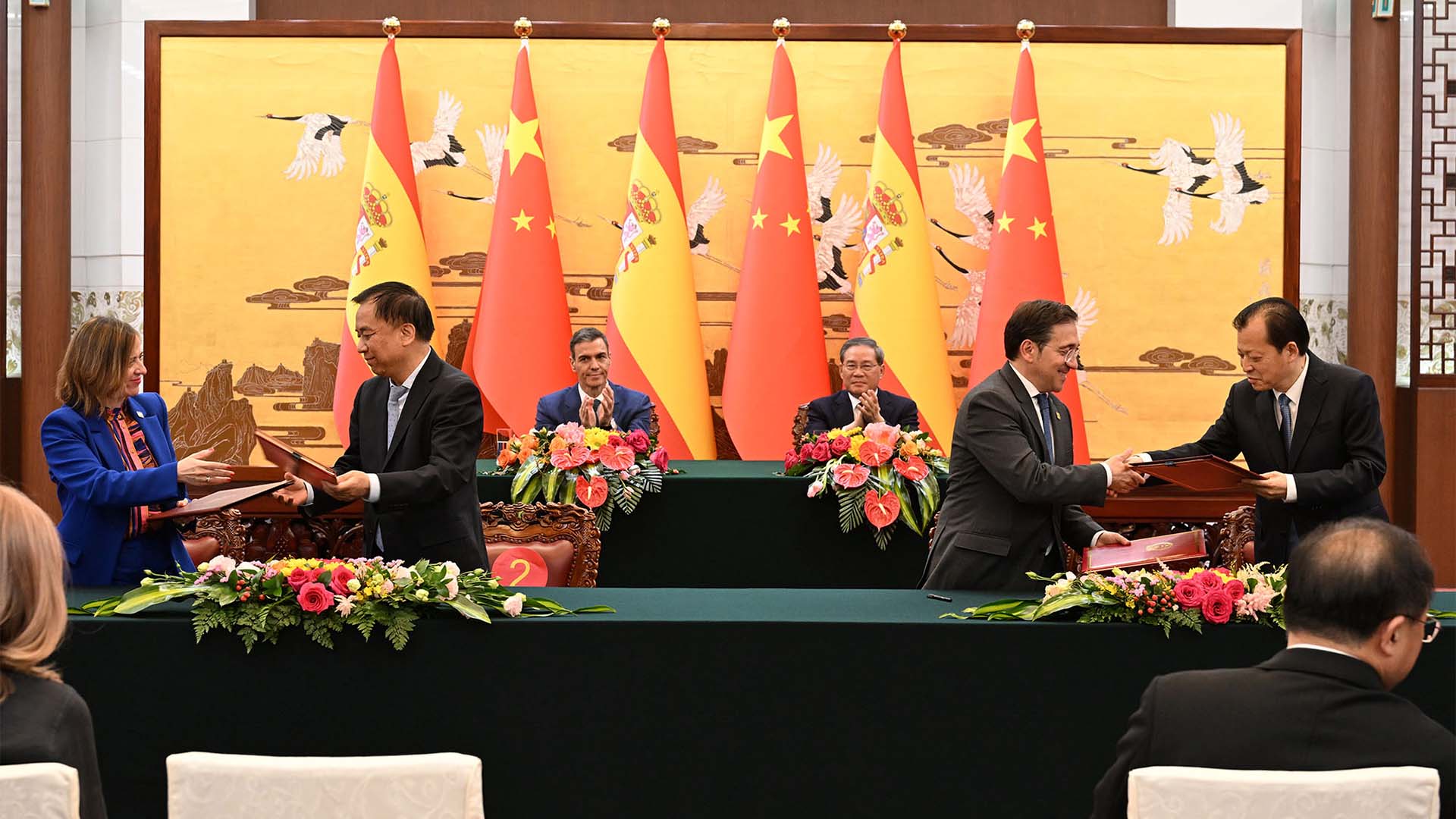 Pedro Sánchez takes part in a signing ceremony in Beijing alongside Chinese Premier Li Qiang, whilst representatives from both governments, including José Manuel Albares, Amparo López Senovilla and Chinese Minister Wang Wentao, exchange documents at an official table with the flags of Spain and China in the background.