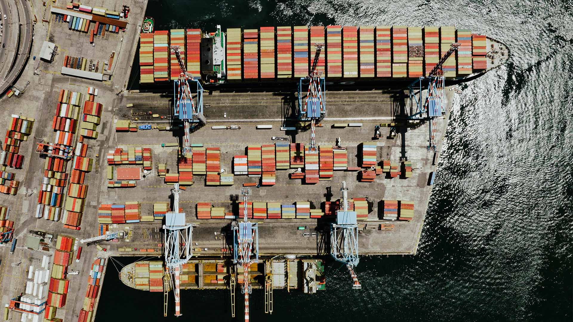 Vista aérea de un puerto marítimo industrial en Nápoles, con varias grúas de carga alineadas y contenedores de transporte multicolores organizados sobre el muelle y a bordo de un buque carguero atracado.