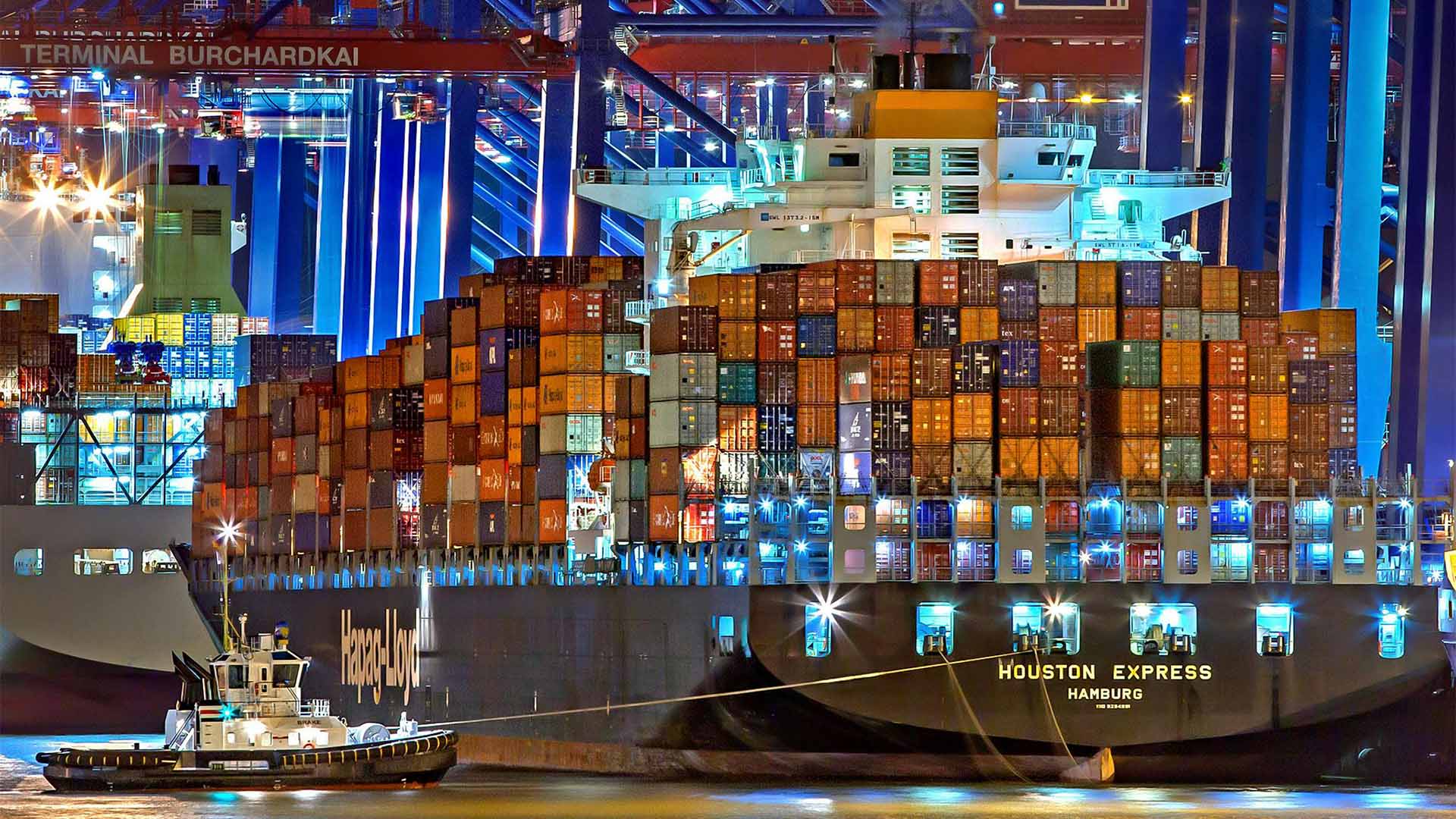 Buque portacontenedores cargado con contenedores de colores en el puerto de Hamburgo, iluminado de noche, con grúas industriales al fondo y un pequeño barco asistiendo en el agua.