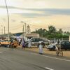 Calle asfaltada en Abiyán al atardecer, con varios taxis amarillos y coches circulando mientras peatones caminan por el arcén y un hombre monta en bicicleta; a la derecha se ven pequeños edificios, un muro con señal policial, postes eléctricos y palmeras.