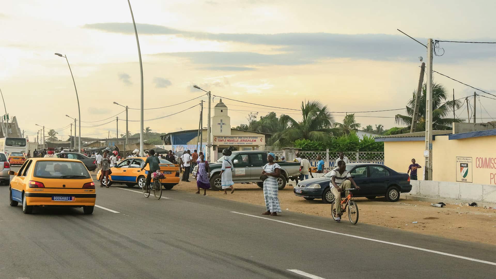 Calle asfaltada en Abiyán al atardecer, con varios taxis amarillos y coches circulando mientras peatones caminan por el arcén y un hombre monta en bicicleta; a la derecha se ven pequeños edificios, un muro con señal policial, postes eléctricos y palmeras.